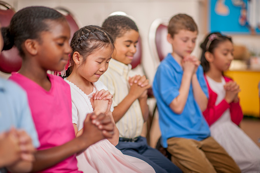 A multi-ethnic group of elementary age children are kneeling on the floor and are praying together.